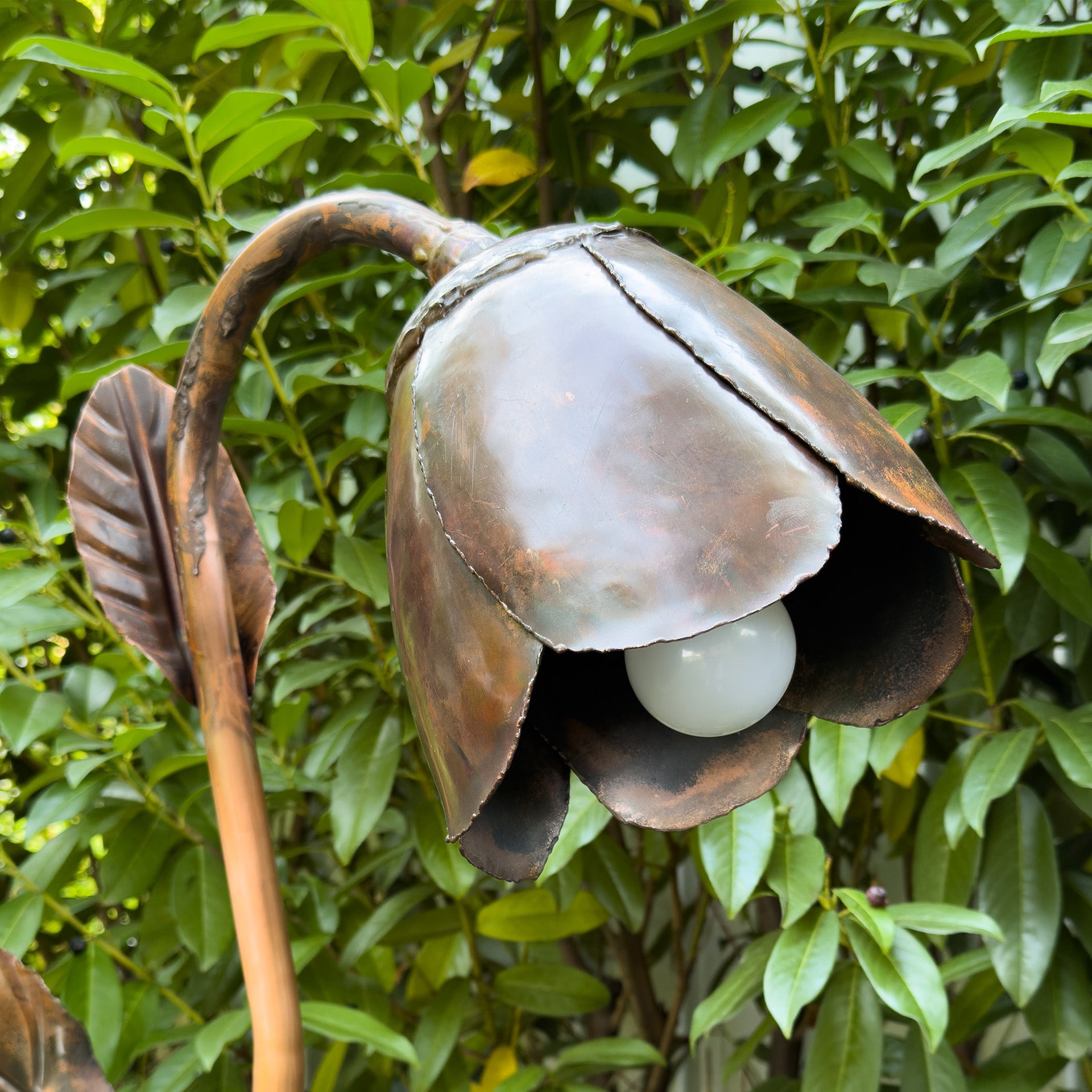 Handgefertigte Kupferlampe in Form einer Blüte mit sichtbarer Glühbirne im Garten.